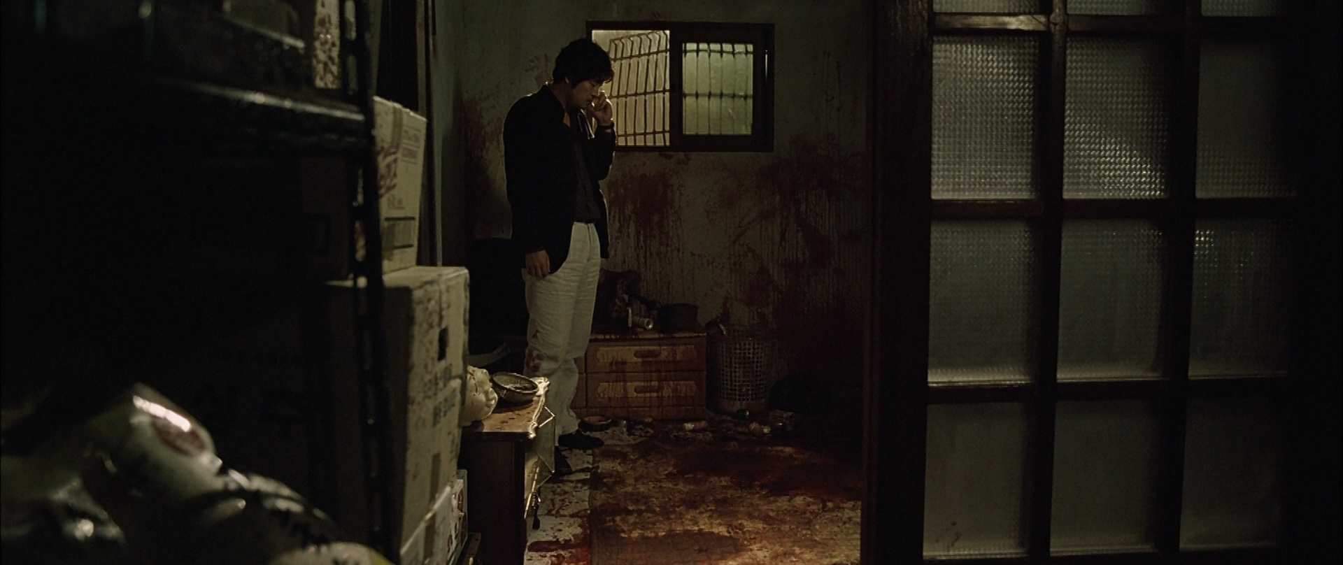 Joong-ho stands in a bloody room while listening to a voicemail on his cellphone.